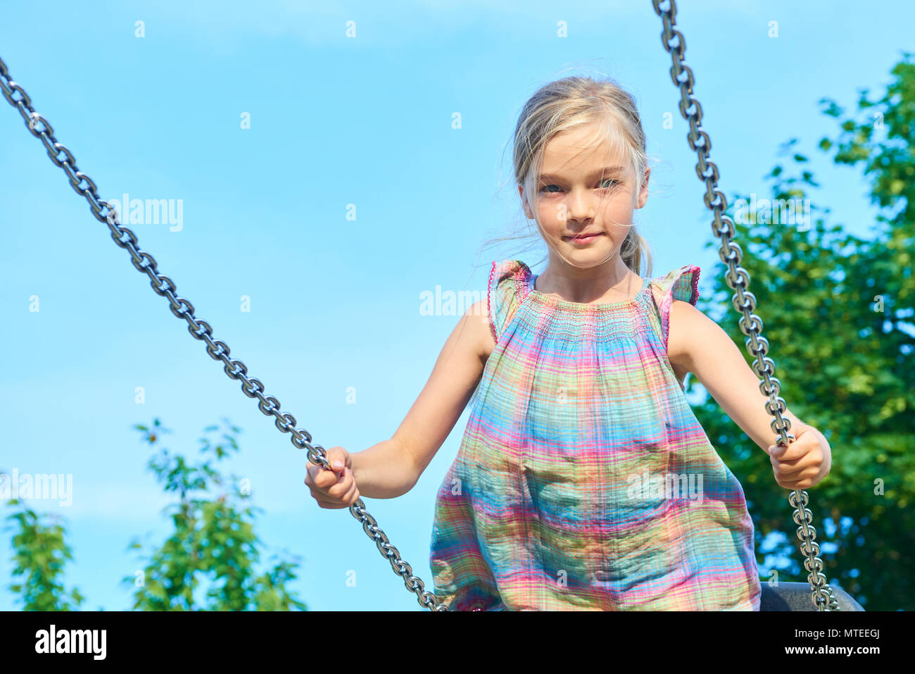 Little child blond girl having fun on a swing outdoor. Summer playground. Girl swinging high. Young child on swing outdoors. Stock Photo