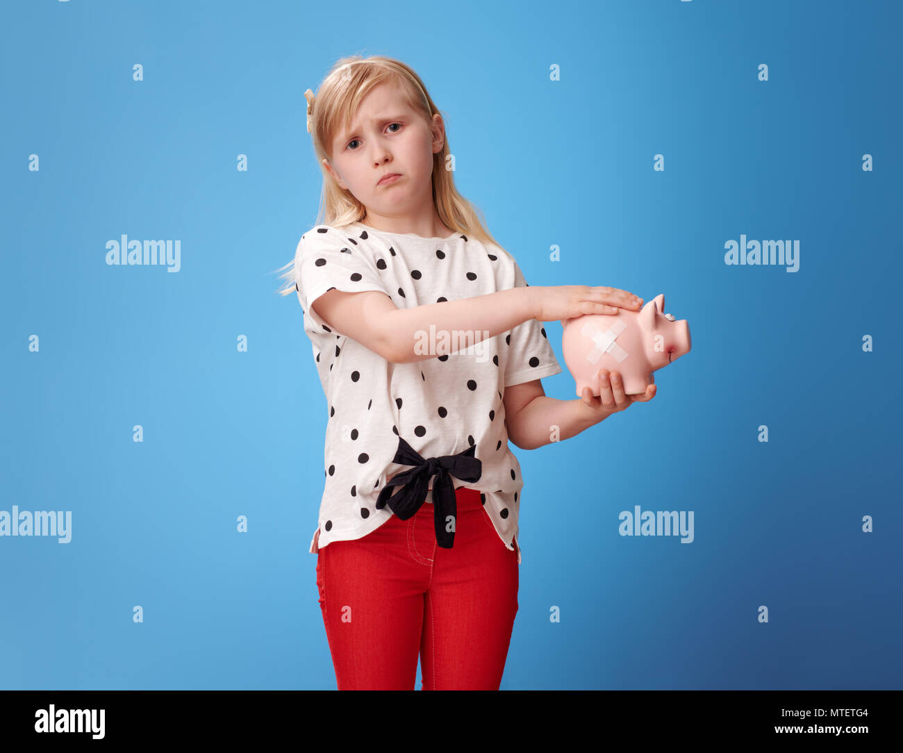 sympathetic modern girl in red pants fondle piggy bank with plaster against blue background Stock Photo