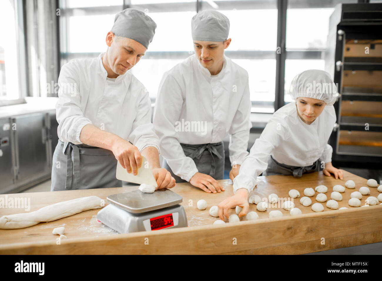 Three young bakers in uniform forming dough for baking on the wooden table standing together at the modern manufacturing Stock Photo