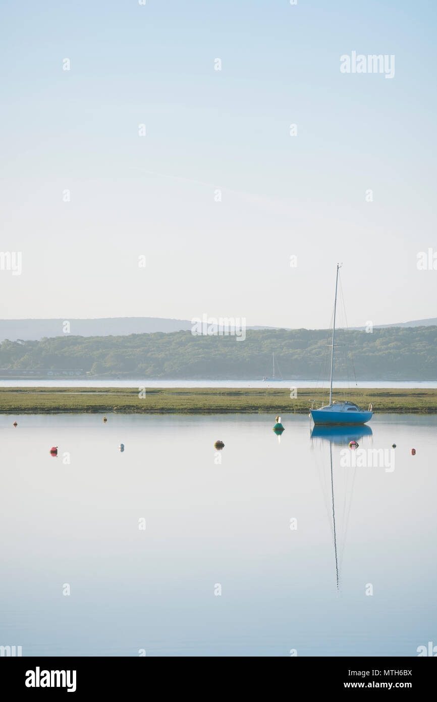 A blue yacht at dawn, moored up and reflected in the calm waters of Keyhaven salt marshes just outside the harbour on a summer morning in June. Stock Photo