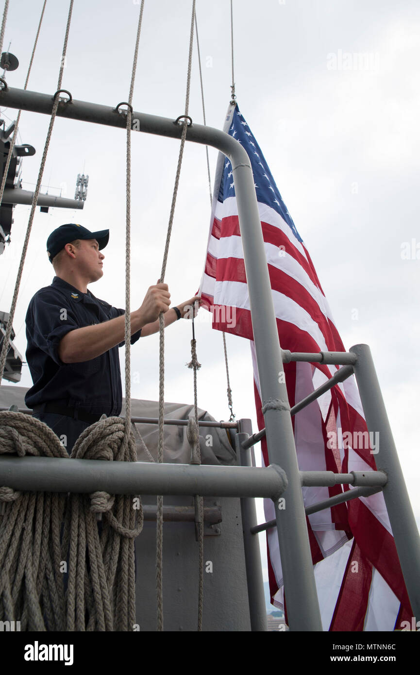 170112-N-XT039-056 SASEBO, Japan (Jan. 12, 2017) Quartermaster 1st Class Matthew Lenerville, from Richardton, N.D., ceremoniously raises an American flag on the main masthead of amphibious assault ship USS Bonhomme Richard (LHD 6) in honor of a U.S. Navy Captain’s retirement. The ceremony is performed at the request of the retiree. The retiree mails the flag to the ship and a flag certificate, signed by the ship’s commanding officer, is sent to the retiree when the flag is returned. Bonhomme Richard, forward-deployed to Sasebo, Japan, is serving forward to provide a rapid-response capability i Stock Photo