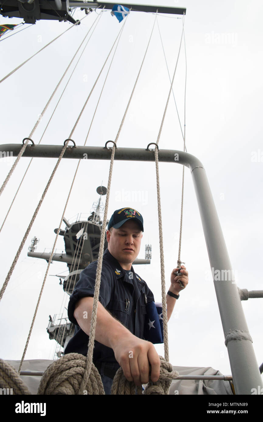 170112-N-XT039-021 SASEBO, Japan (Jan. 12, 2017) Quartermaster 1st Class Matthew Lenerville, from Richardton, N.D., unties a halyard aboard amphibious assault ship USS Bonhomme Richard (LHD 6) prior to raising an American flag in honor of a U.S. Navy Captain’s retirement. The ceremony is performed at the request of the retiree. The retiree mails the flag to the ship and a flag certificate, signed by the ship’s commanding officer, is sent to the retiree when the flag is returned. Bonhomme Richard, forward-deployed to Sasebo, Japan, is serving forward to provide a rapid-response capability in th Stock Photo