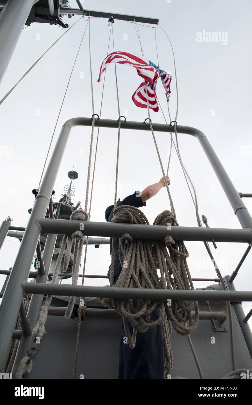 170112-N-XT039-072 SASEBO, Japan (Jan. 12, 2017) Quartermaster 1st Class Matthew Lenerville, from Richardton, N.D., ceremoniously raises an American flag on the main masthead of amphibious assault ship USS Bonhomme Richard (LHD 6) in honor of a U.S. Navy Captain’s retirement. The ceremony is performed at the request of the retiree. The retiree mails the flag to the ship and a flag certificate, signed by the ship’s commanding officer, is sent to the retiree when the flag is returned. Bonhomme Richard, forward-deployed to Sasebo, Japan, is serving forward to provide a rapid-response capability i Stock Photo