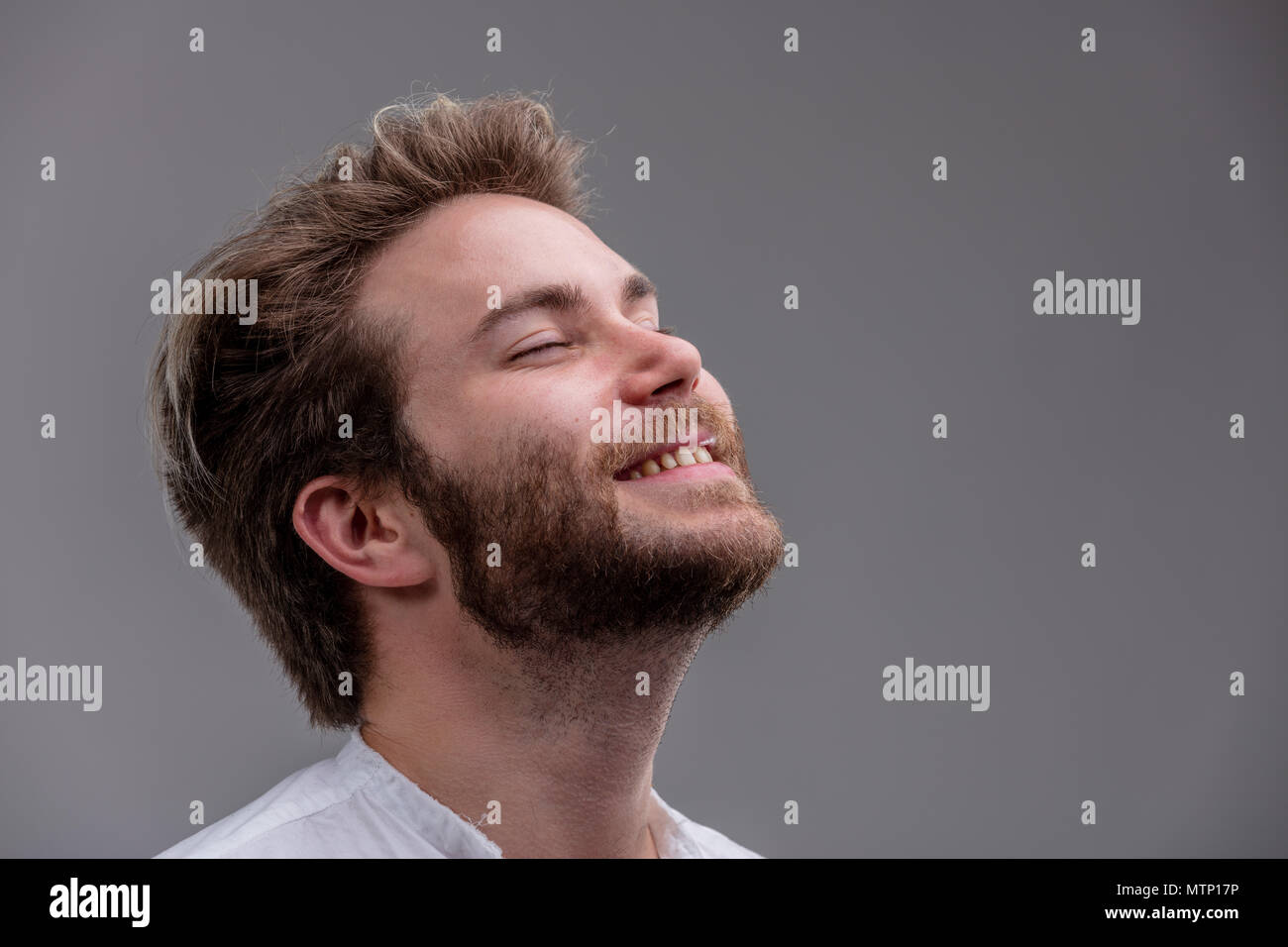 Blissful relaxed bearded young man with his head tilted back, closed eyes and a beaming smile isolated on grey with copy space Stock Photo