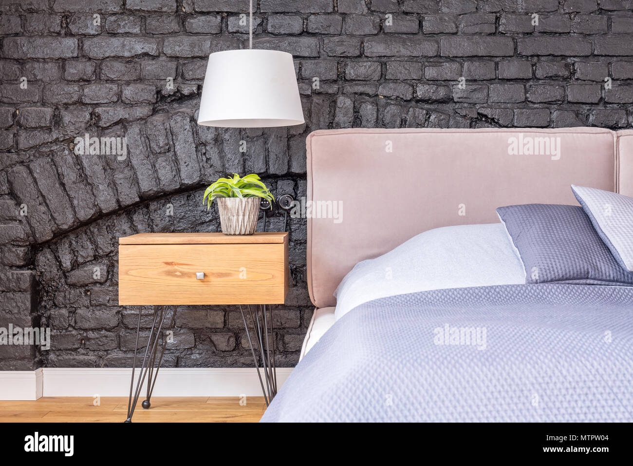 Small plant in pot on wooden bedside cabinet under white lamp in bedroom with brick wall Stock Photo