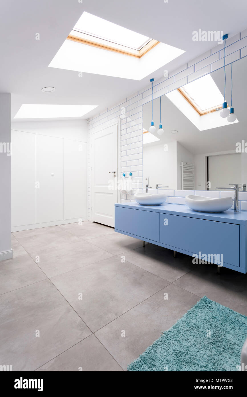 Cozy attic bathroom with grey floor, blue washbasin cabinet, mirror and window Stock Photo