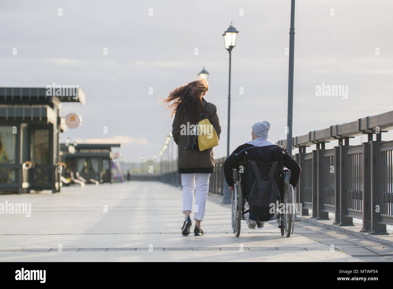 Rear view of young woman walking with disabled man in wheelchair on the ...