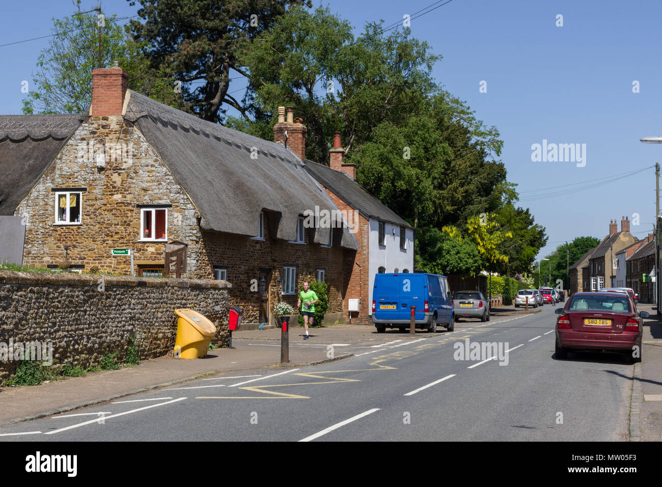 The main street through the village of Little Harrowden, with a fine thatched property to the left plus a lone jogger; Northamptonshire, UK Stock Photo