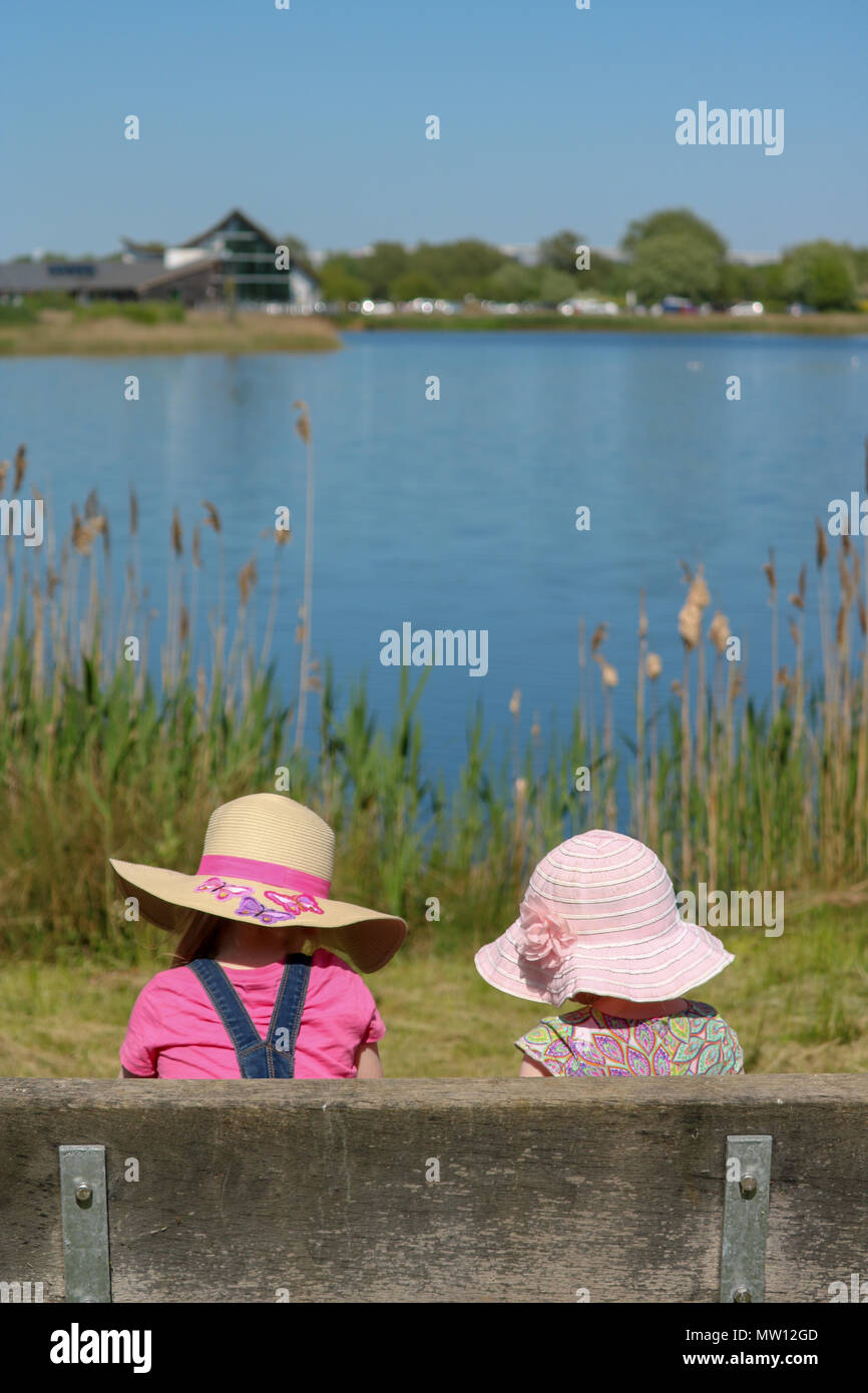 A young girl and a toddler, backs to camera and both in sunhats, sit on a bench and contemplate the view over a lake on a gloriously sunny day at Stanwick Lakes, Northampton, UK Stock Photo