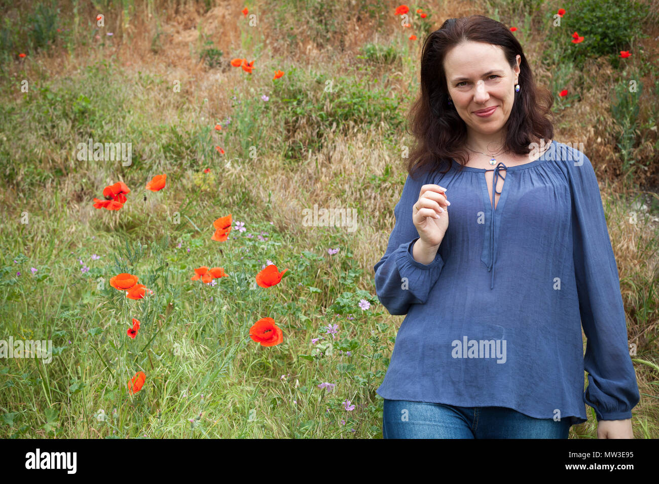 Smiling young adult European woman walks on a summer meadow with flowering poppies. Outdoor portrait Stock Photo