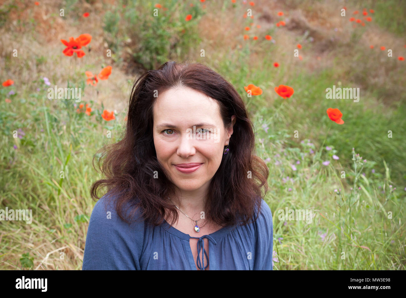 Outdoor portrait of smiling young European woman rests on a summer meadow with flowering poppies Stock Photo