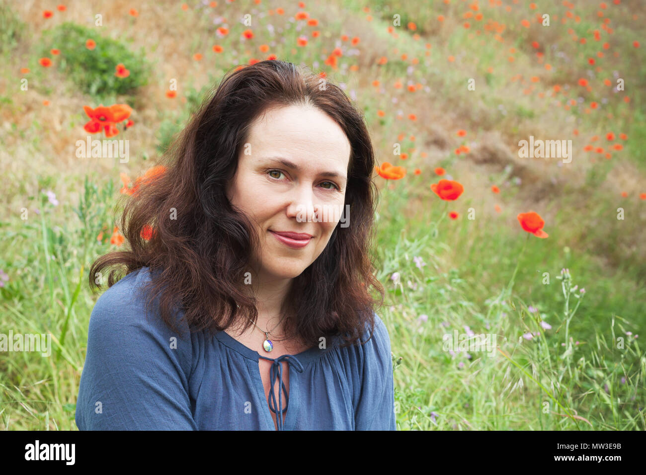 Smiling young adult European woman rests on a summer meadow with flowering poppies. Outdoor portrait Stock Photo