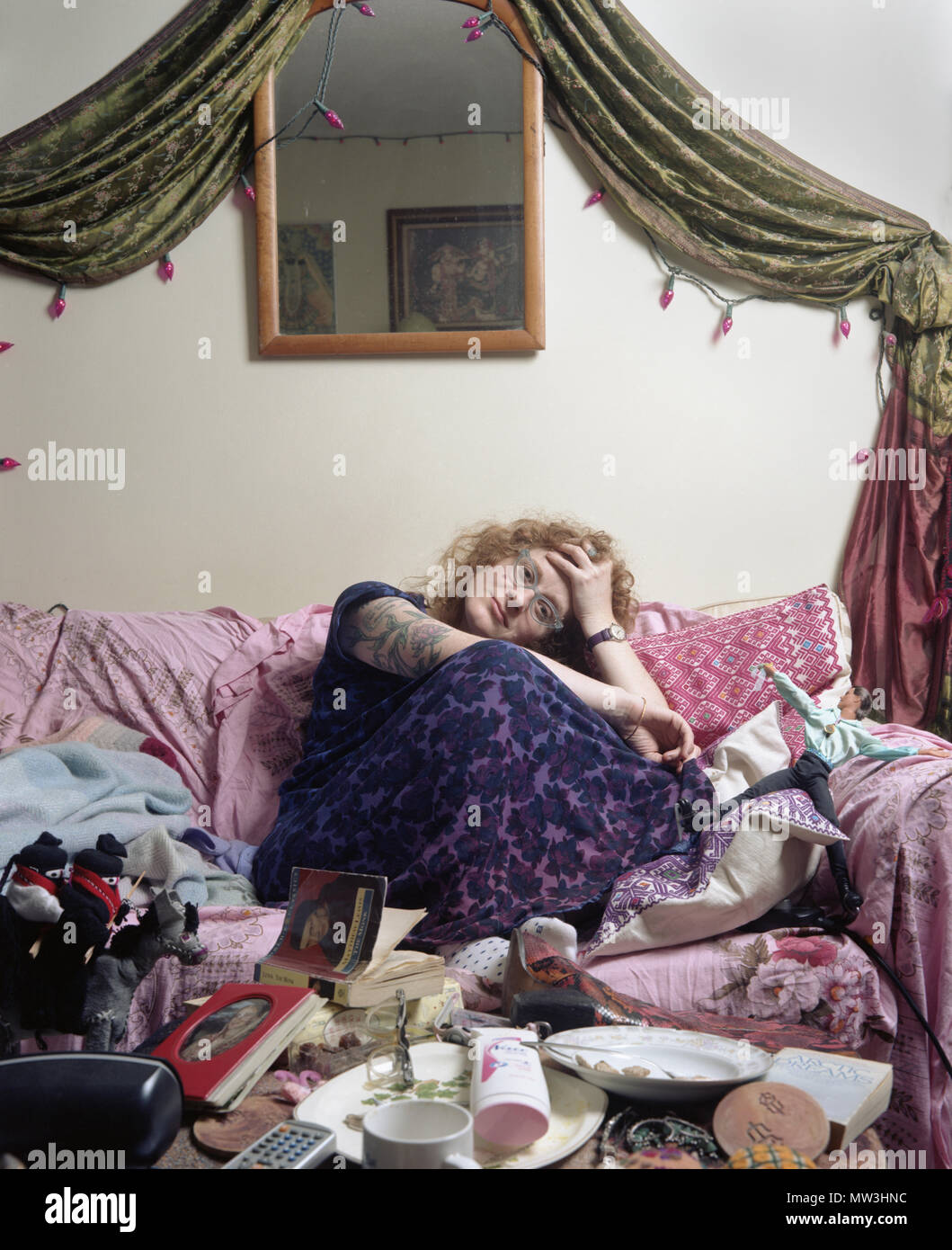 Tattooed woman in purple nightgown sitting in a cluttered living room gazing peacefully into the camera. Stock Photo