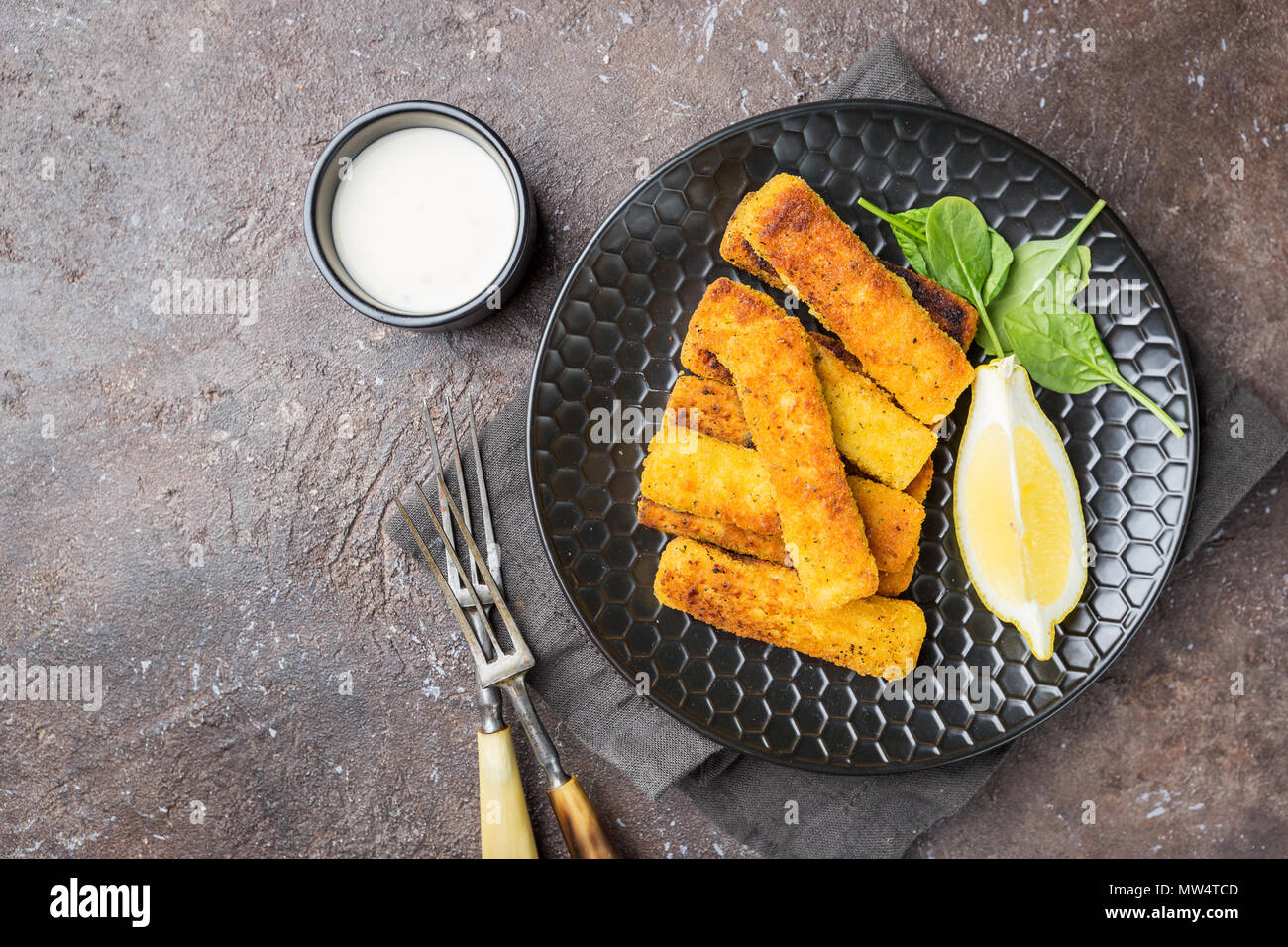 Tasty savory snack of crumbed fish fingers sticks served on a plate with lemon over dark stone background, top view with copy space Stock Photo