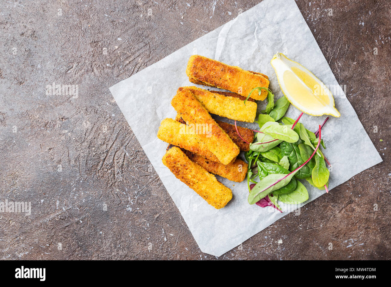 Tasty savory snack of crumbed fish fingers sticks served on paper with lemon over dark stone background, top view with copy space Stock Photo