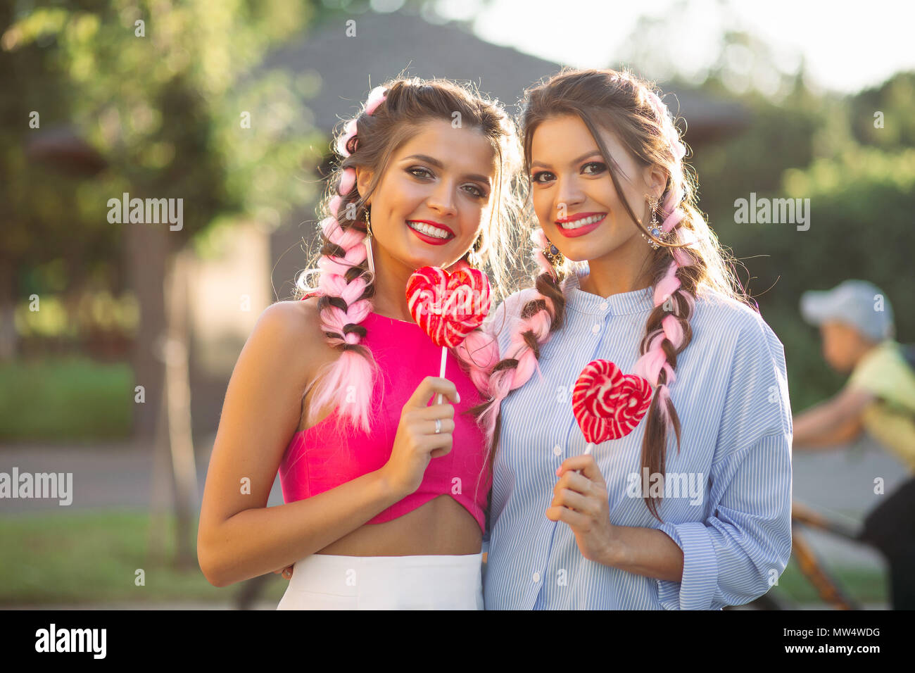 Girlfriends with colorful braids holding candy heart on stick. Stock Photo