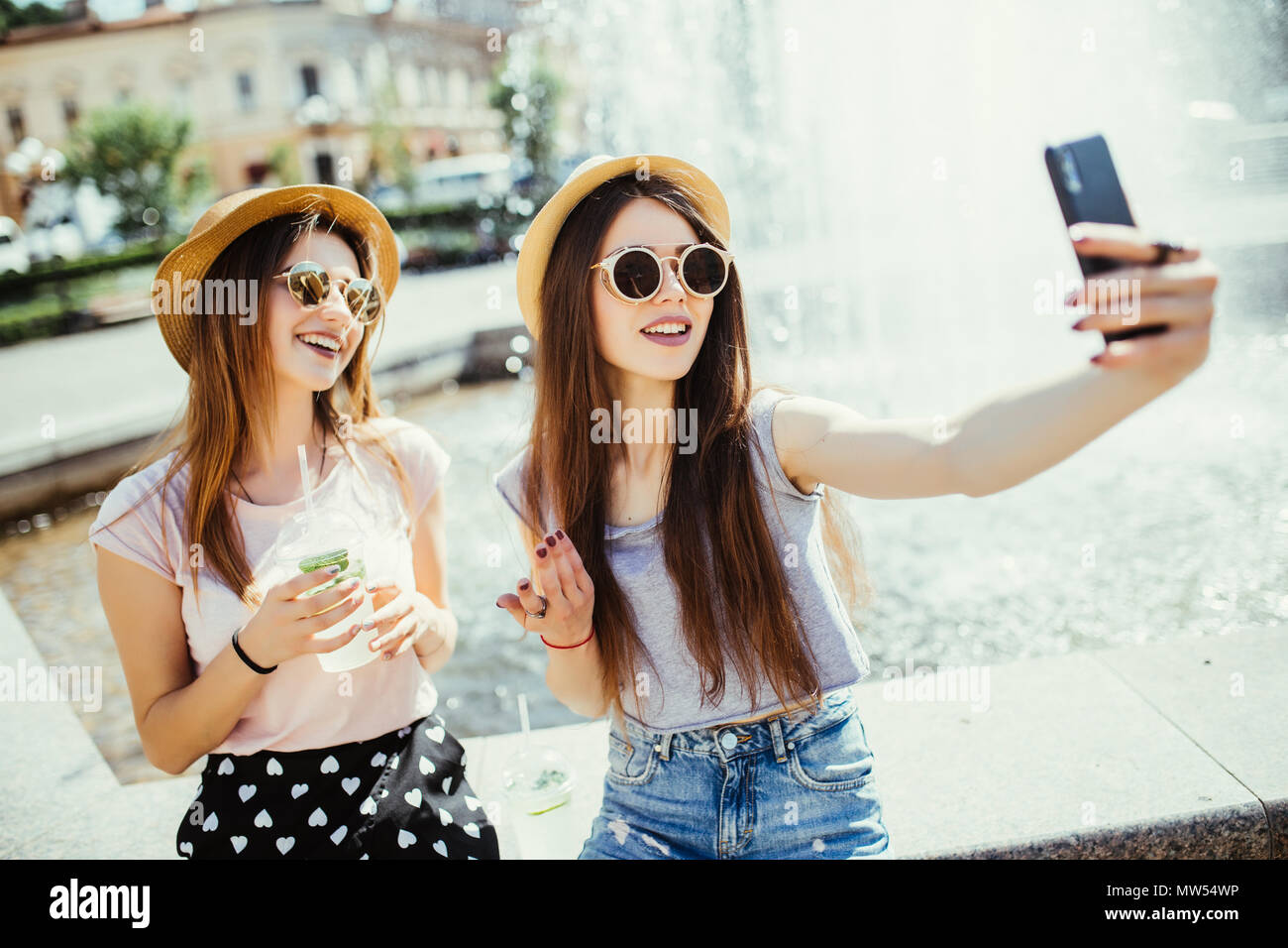 Two female best friends recreat together against fountain, look cheerfully at modern smart phone, message with friends, connected to wireless internet Stock Photo