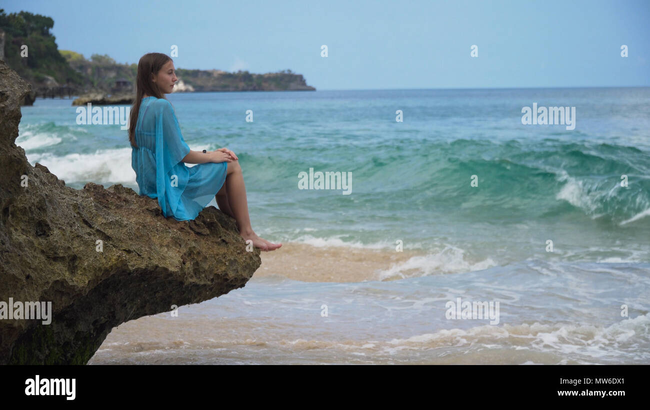 Beautiful girl in blue dress sitting on a rock above the sea and ...