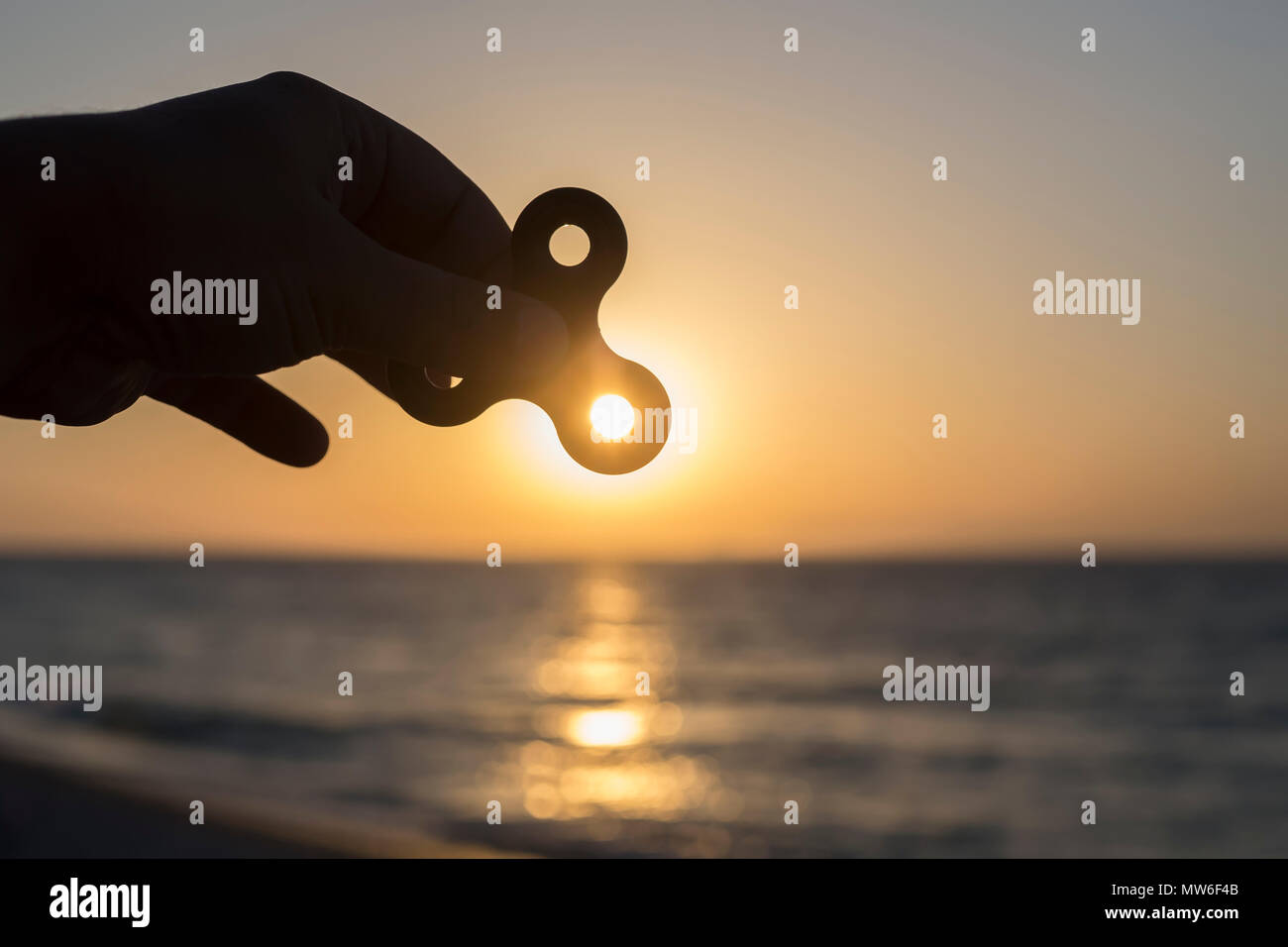 Siluete of hand holding fidget spinner at sunset on the sea, stress relieving toy. Stock Photo