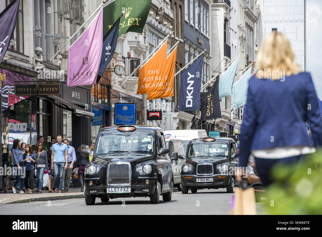 A busy day in Mayfair's up market retail district. A women with shopping bags walks as black cabs drive down the street lined with branded flags Stock Photo