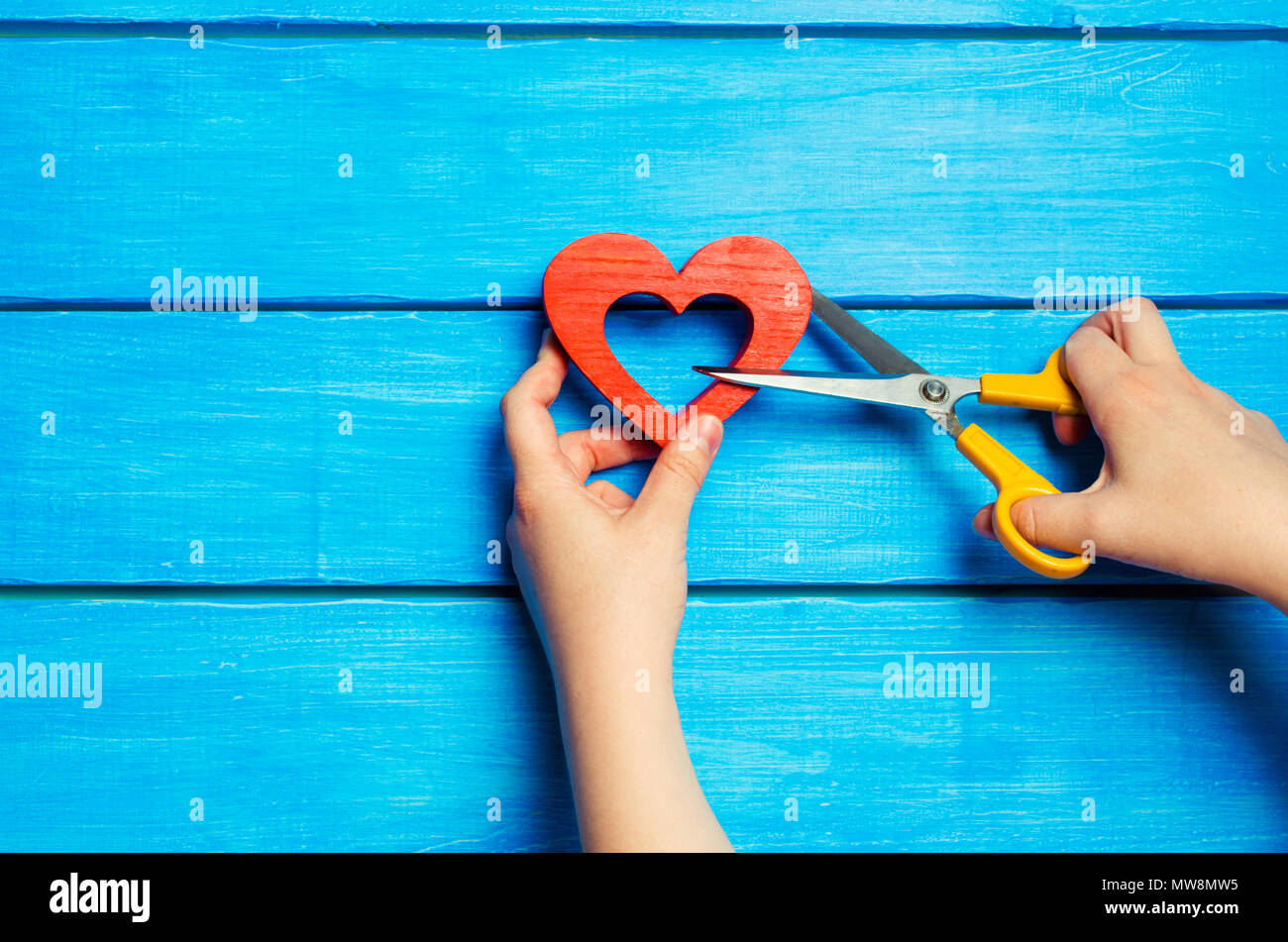 Female hands cut a wooden red heart with scissors on a blue background. The concept of breaking relations, quarrel, divorce. The pair diverges. Betray Stock Photo