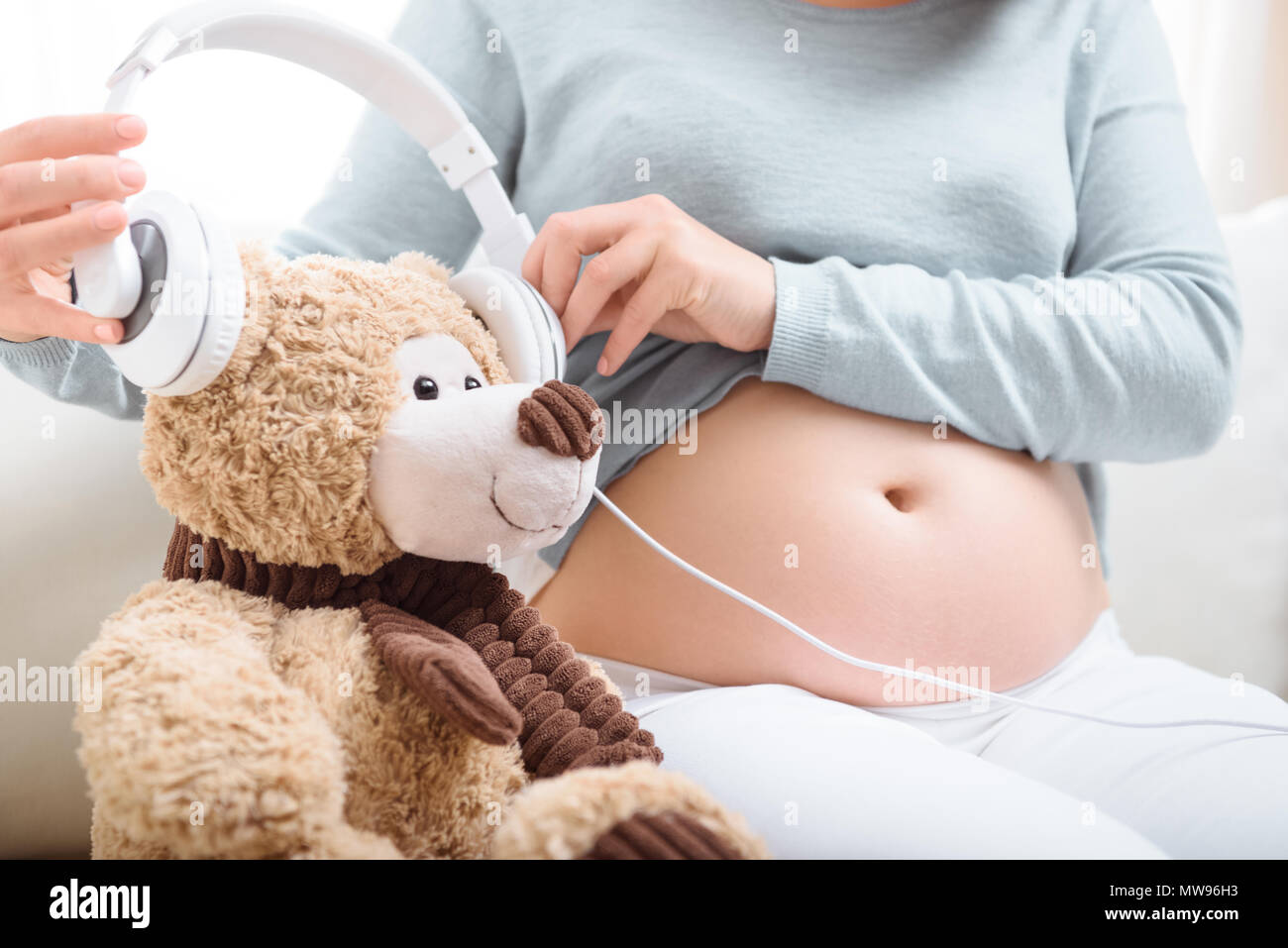 charming redhead pregnant woman with headphones on her belly sitting on  sofa with teddy bear, Stock image