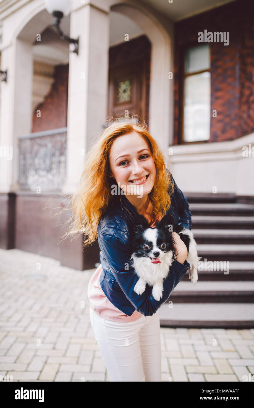 A beautiful young woman with red long hair is holding a small, cute funny big-eyed dog of two flowers, a black-and-white pet of the breed of hichuahua Stock Photo