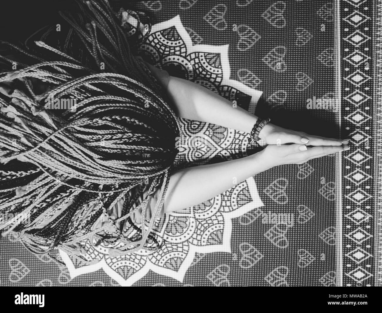 Concentrated woman with blue african braids praying with wooden rosary mala beads. Namaste. Close up hands on yoga mat with mandala. Top view. Black and white Stock Photo