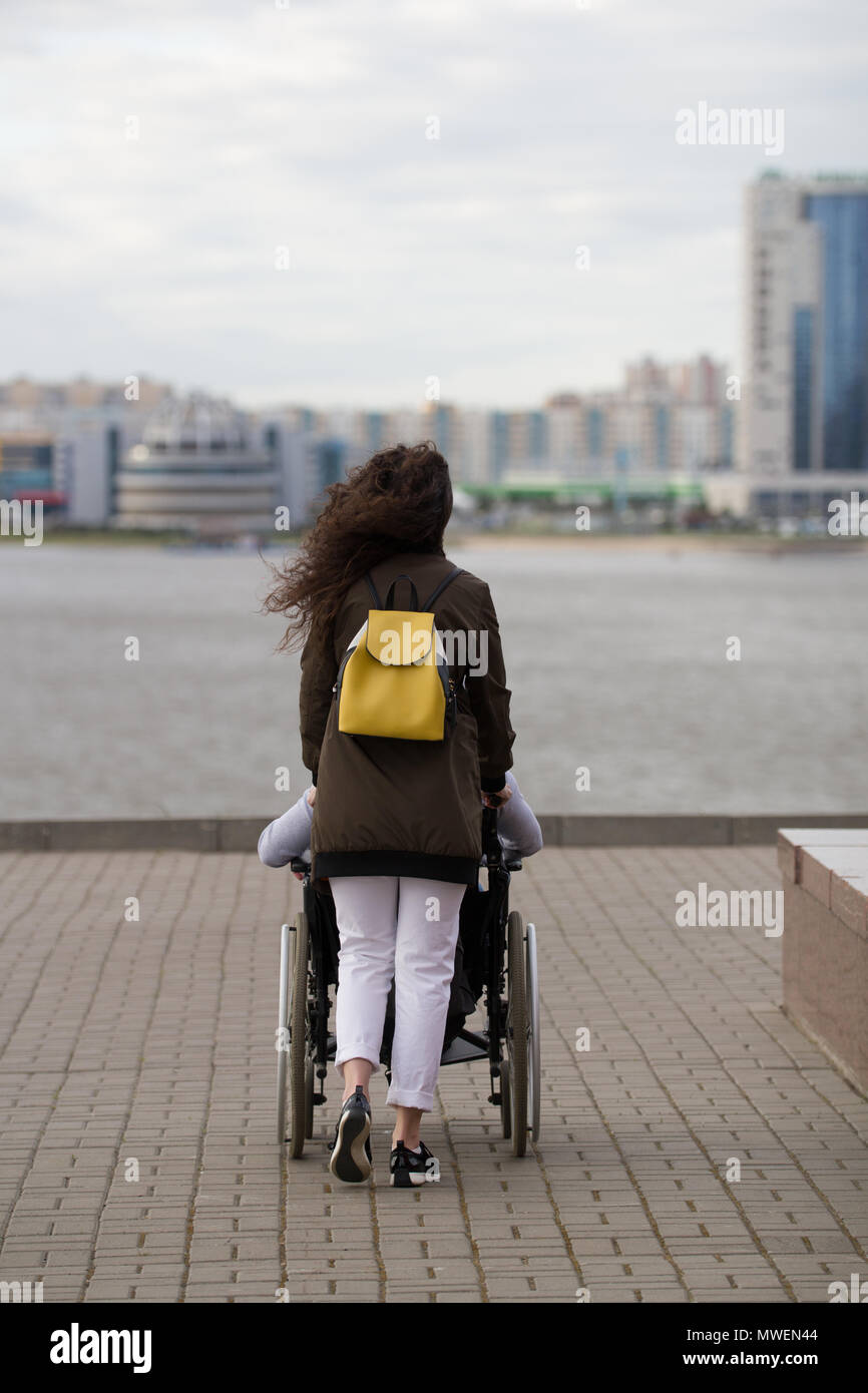 Rear view of young woman walking with disabled man in wheelchair on the ...