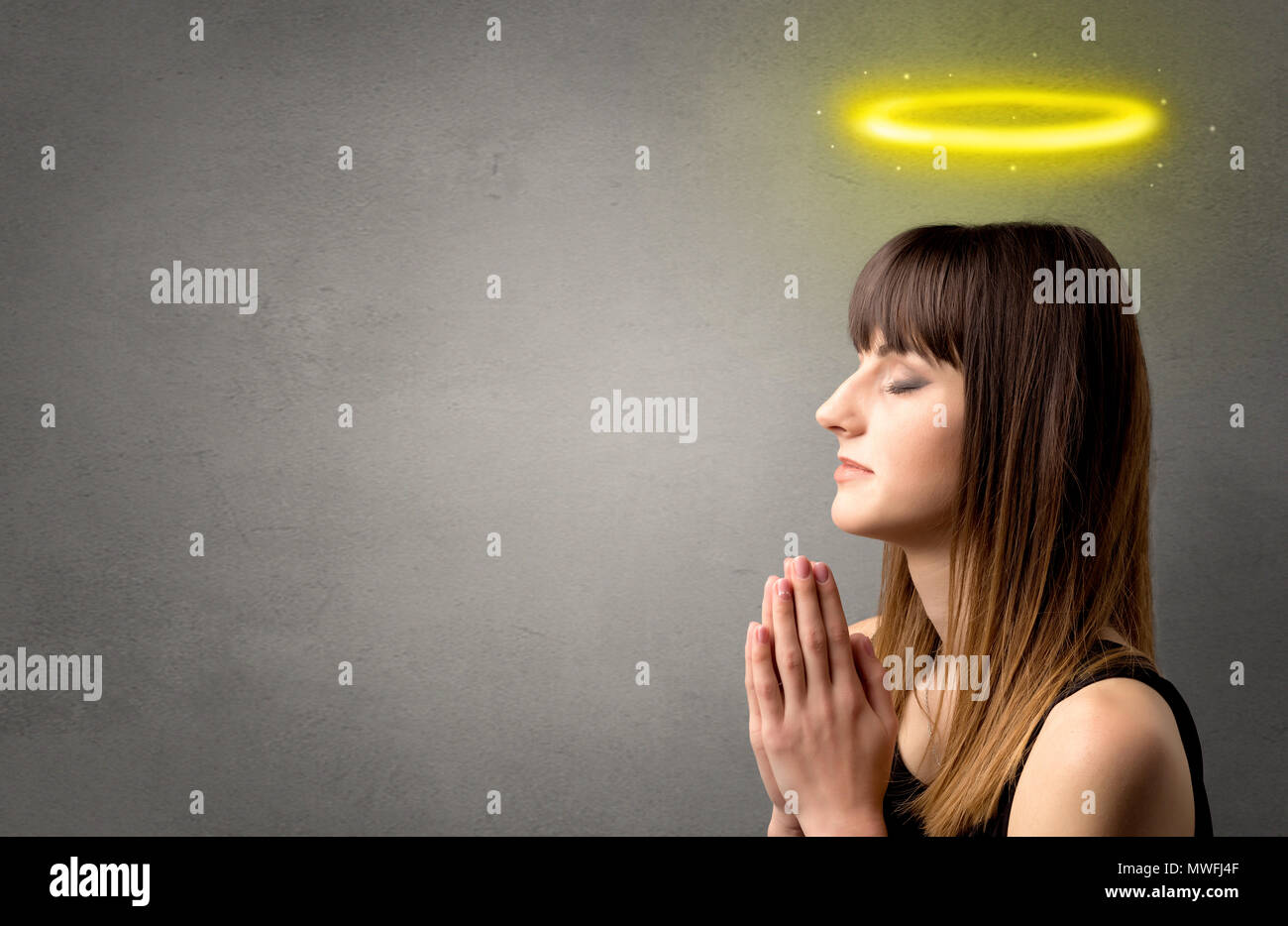 Young woman praying on a grey background with a shiny yellow halo above ...