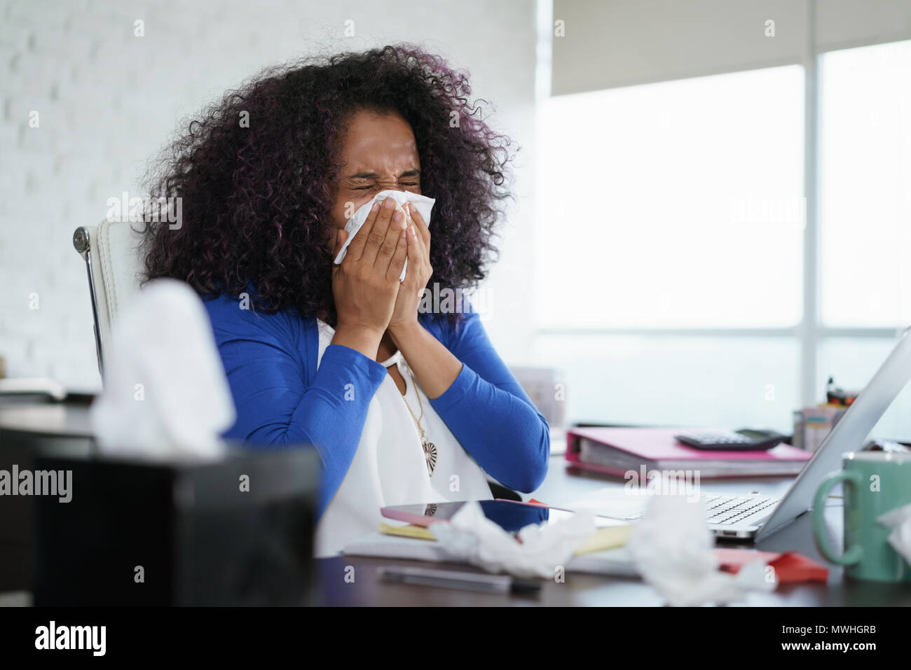 Sick african american girl working from home office. Ill young black woman with cold, sitting at desk with laptop computer and sneezing for allergy. Stock Photo