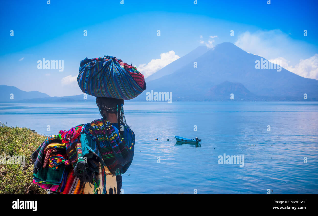 Panajachel, Guatemala -April, 25, 2018: Outdoor view of unidentifed indigenous woman, wearing typical clothes and walking in lakeshore with small boats in Atitlan Lake and volcano in Background in Guatemala Stock Photo
