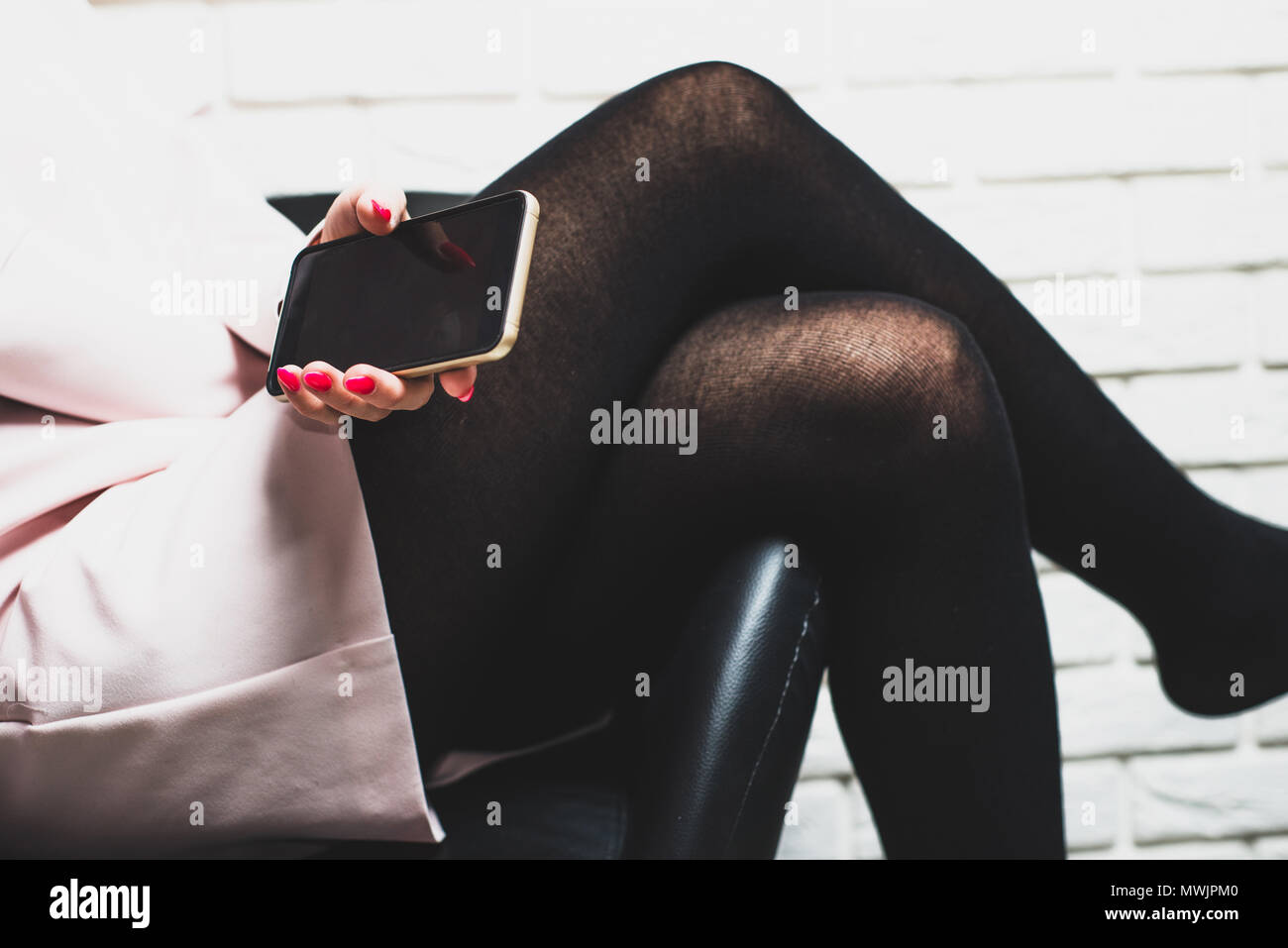 A woman sitting on a chair holds in her hand a phone directed to the camera. A woman wearing a pink skirt and black tights sits worried, thoughtful, p Stock Photo