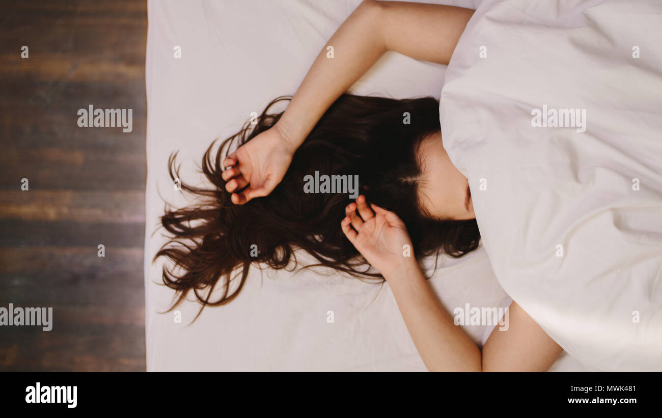 Top view of a woman sleeping on bed covering her face. Close up of a woman in deep sleep with her hand beside her head and hair let loose. Stock Photo