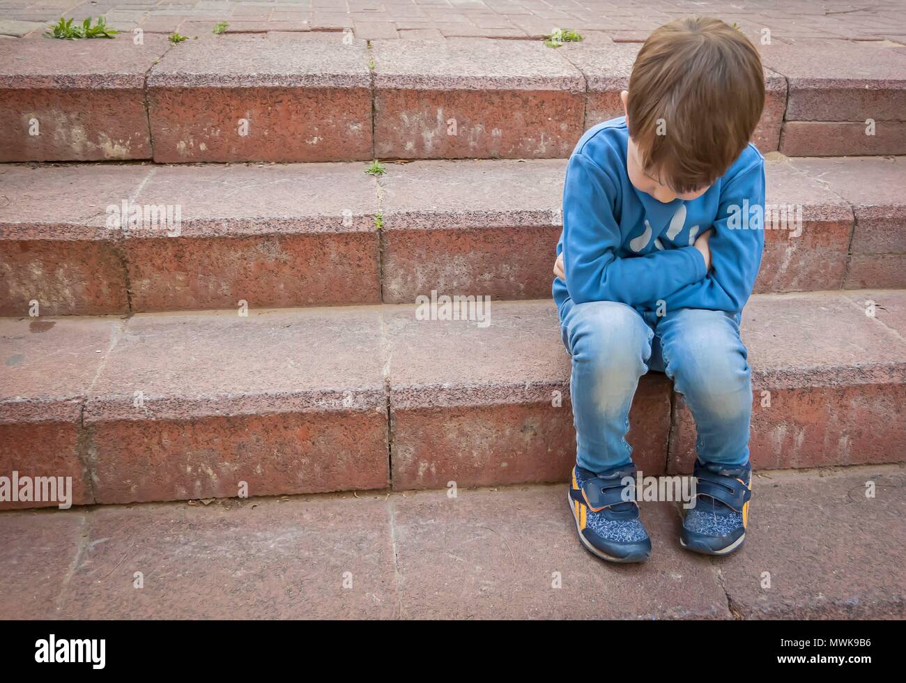 Unhappy little Caucasian child sitting on the stairs in a closed position with his head down. Illustrative image for social issues, child offense Stock Photo