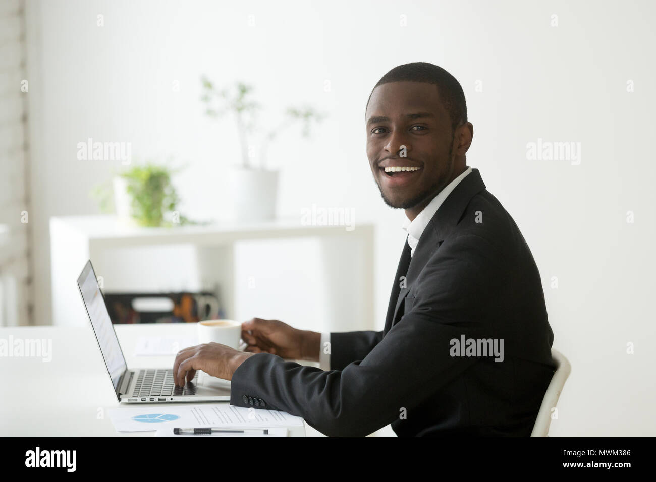 Smiling black worker working at laptop and drinking coffee Stock Photo