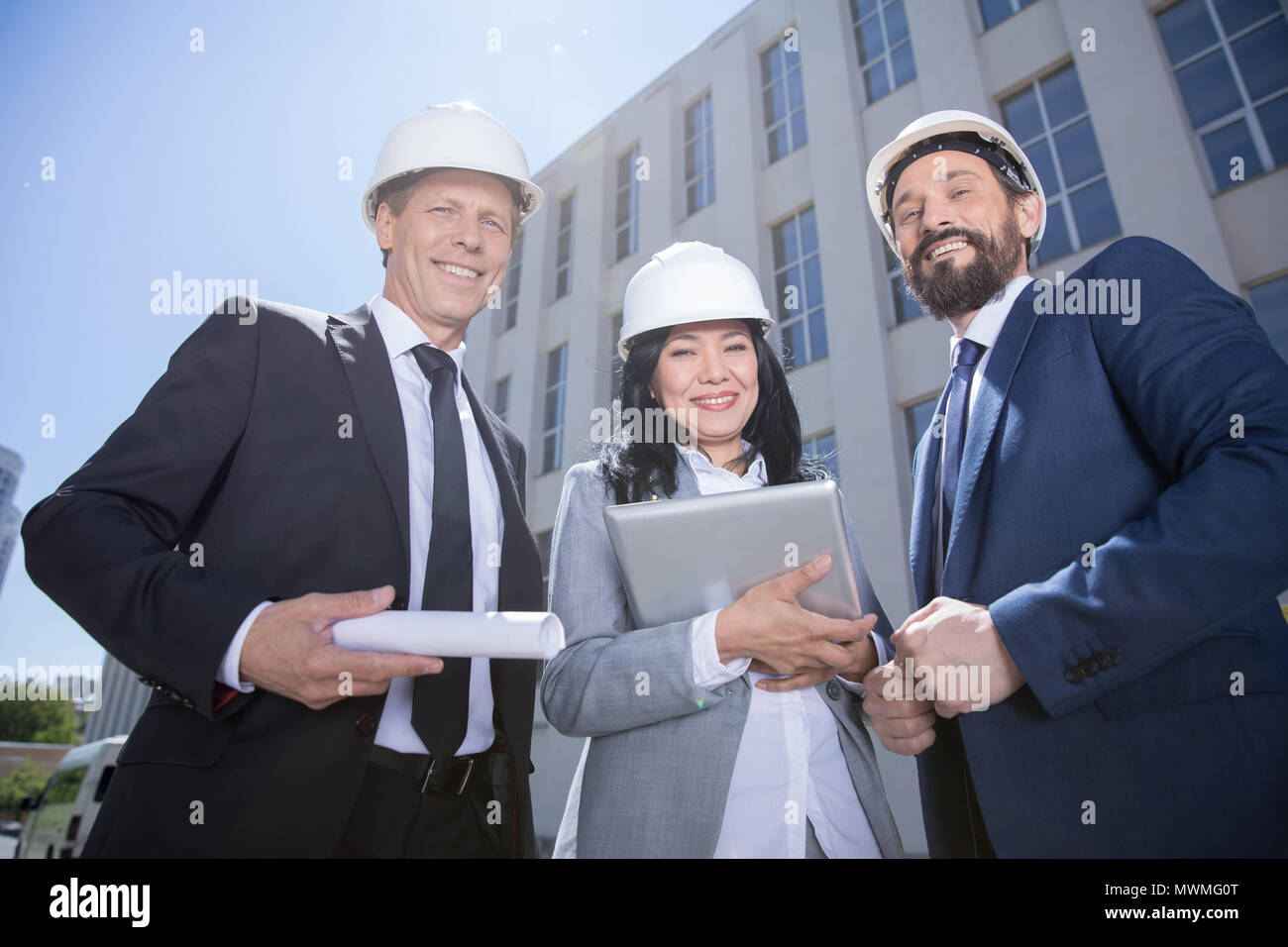 Smiling professional architects in hard hats holding blueprint and using digital tablet Stock Photo