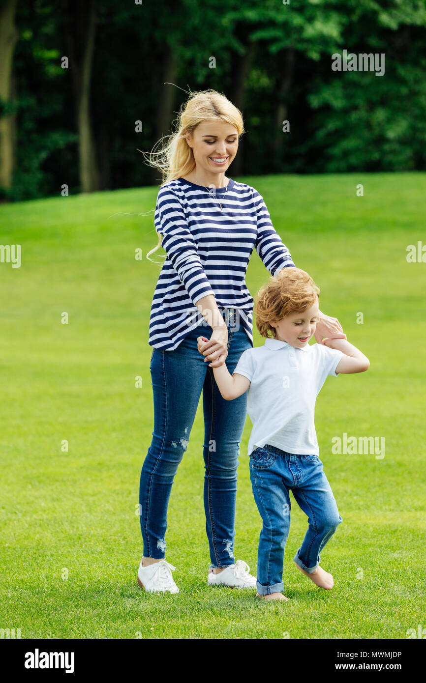 happy young mother with cute little son holding hands while walking on green lawn in park Stock Photo