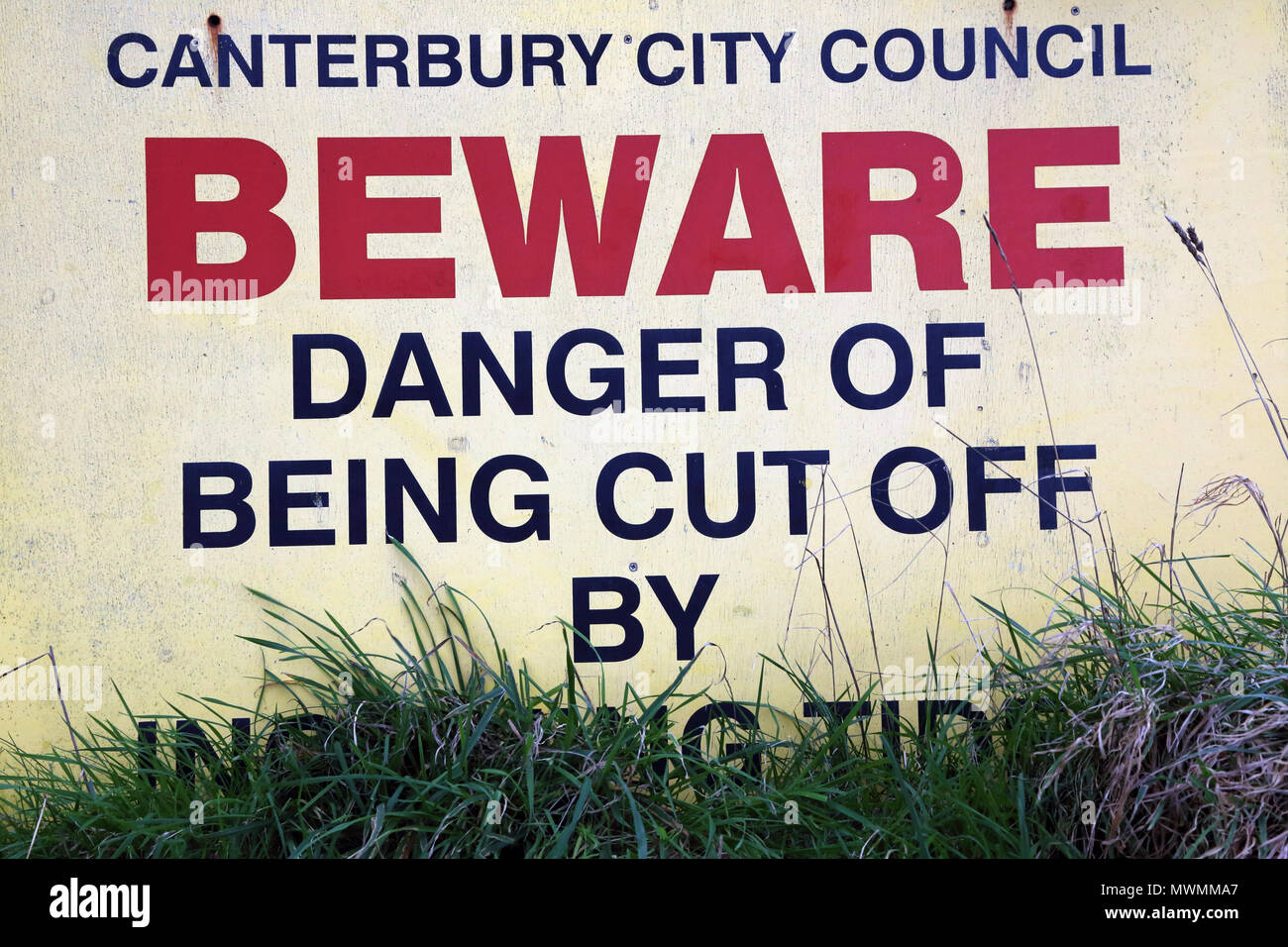 Sign warning of the danger of being cut off by incoming tide, partially obscured by grass. Near Herne Bay, Kent, UK. Stock Photo