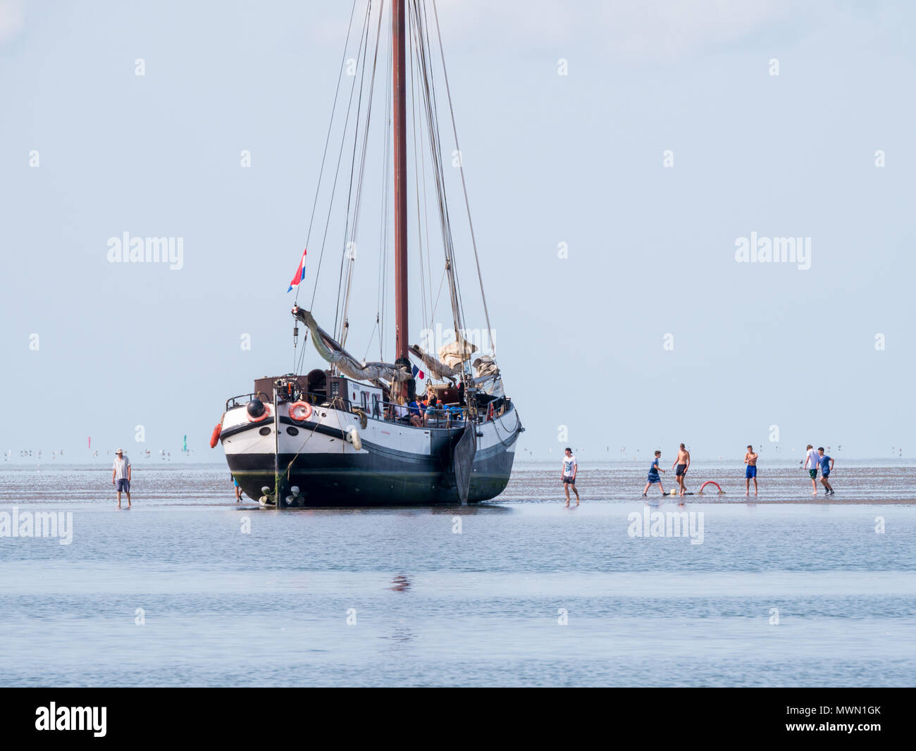 Group of youngsters playing on sand flat and dried out flat-bottom sailing yacht at low tide on Wadden sea, Netherlands Stock Photo