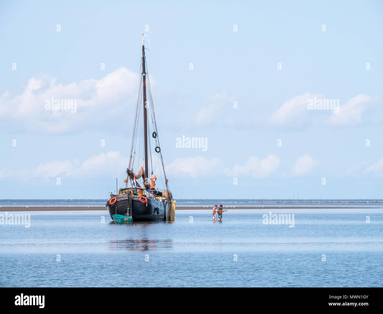 Young men wading in shallow water near sand flat and sailing yacht aground at low tide on Wadden sea, Netherlands Stock Photo