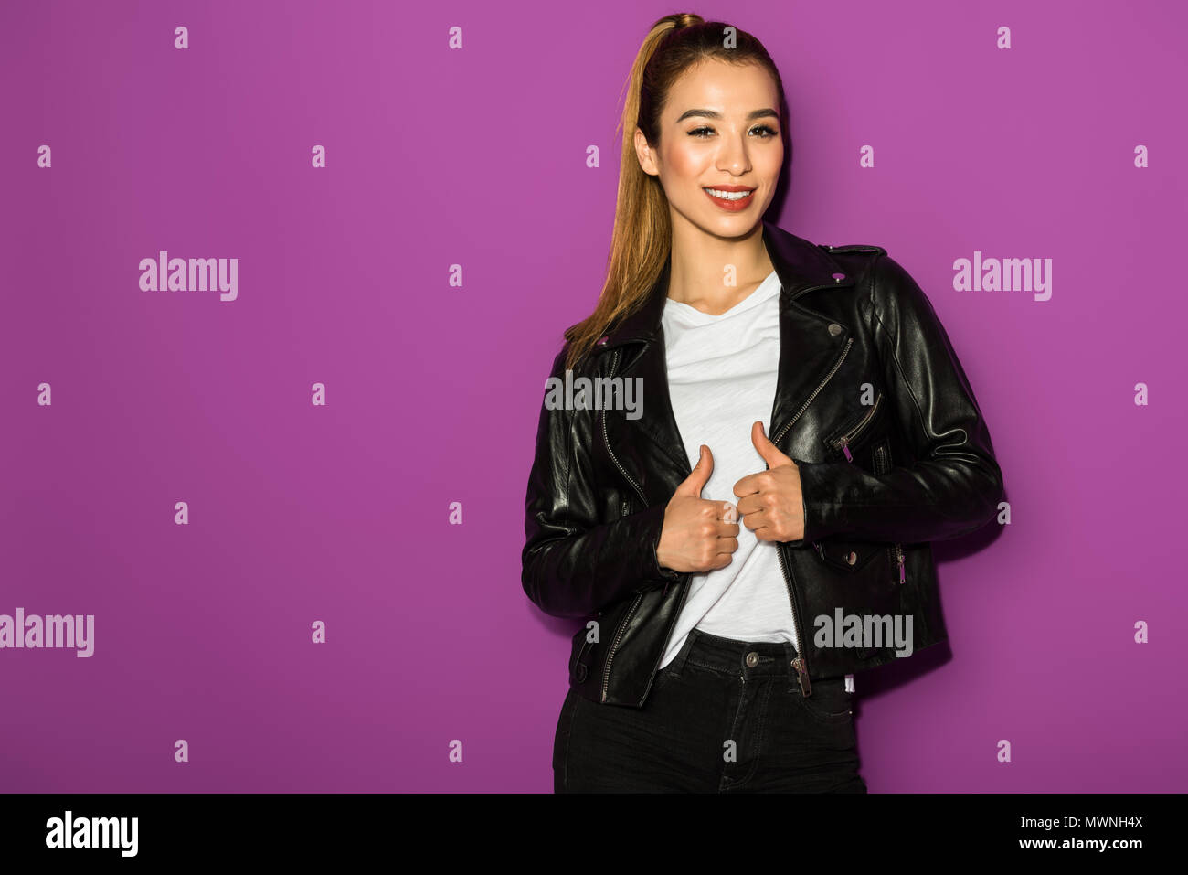 beautiful stylish asian girl in leather jacket smiling at camera isolated on violet Stock Photo