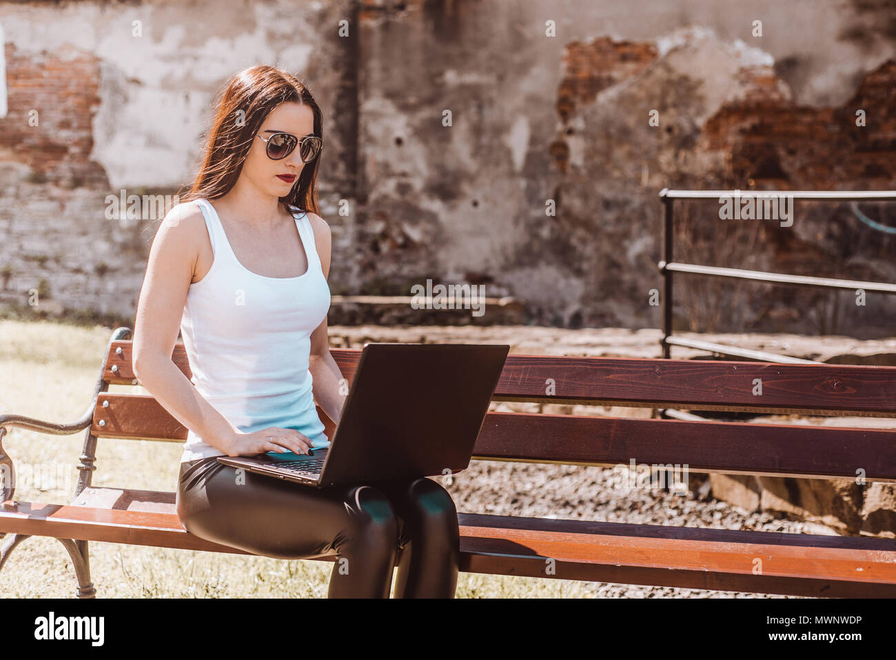 Girl sits on a bench outside with a laptop and works. A woman in sunglasses transfers work to fresh air. Relaxation and work. Stock Photo
