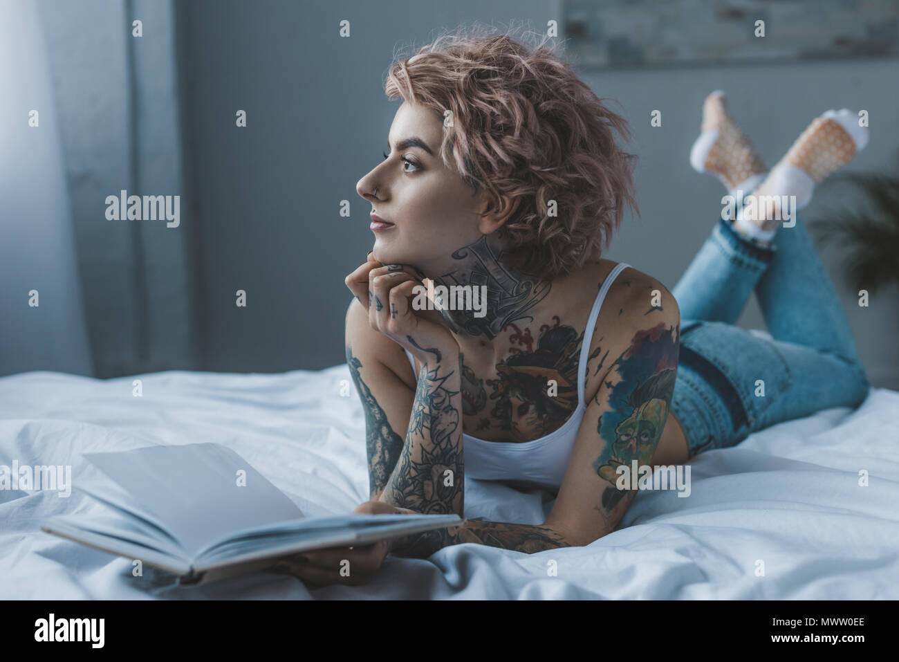 thoughtful tattooed girl lying on bed with book Stock Photo
