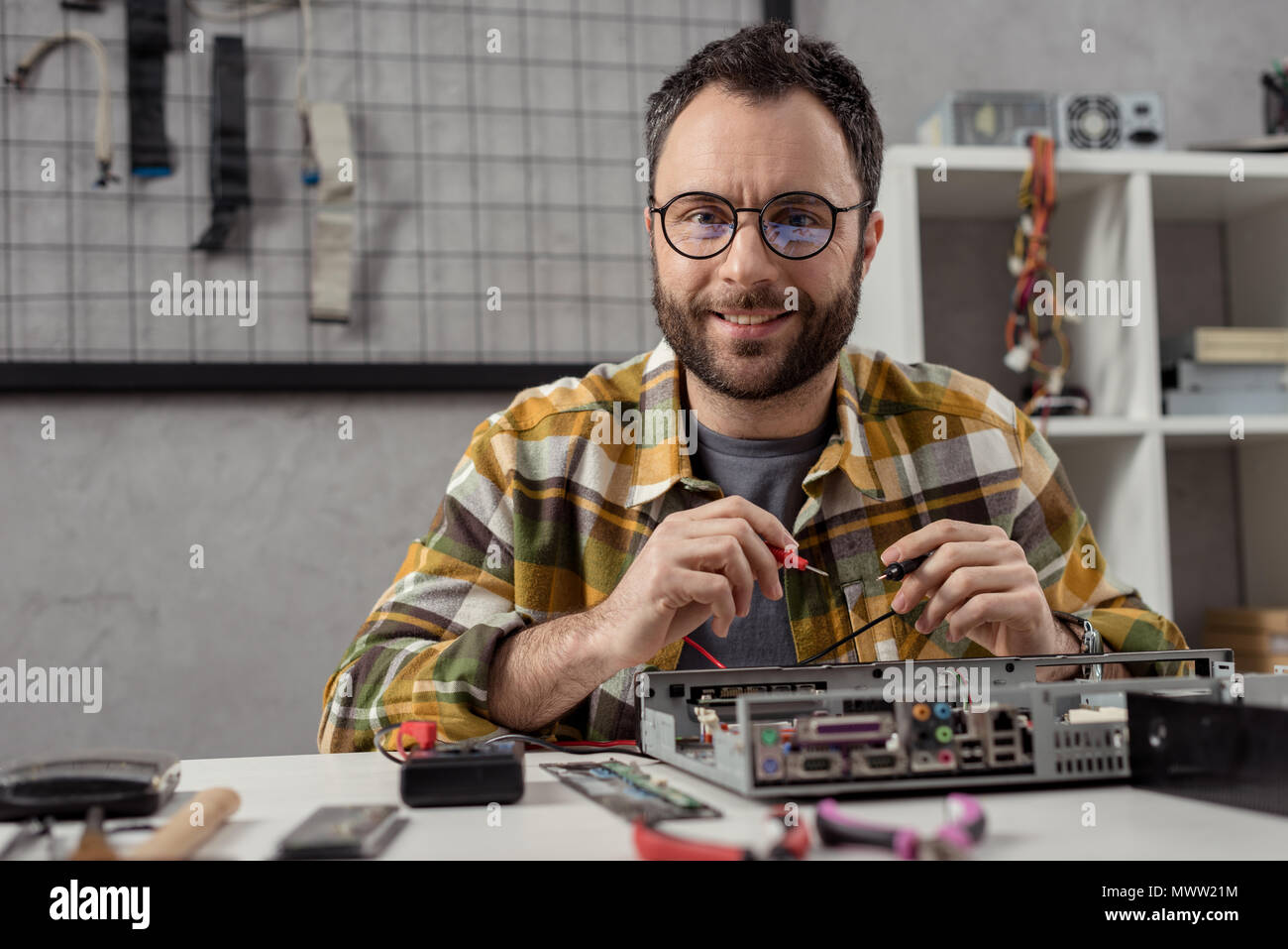 smiling man looking at camera while using multimeter over broken pc Stock Photo