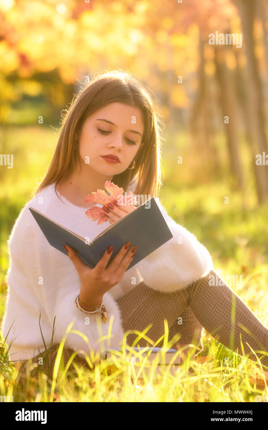 Girl reading a book sitting in the nature with a fallen leaf in her hand Stock Photo