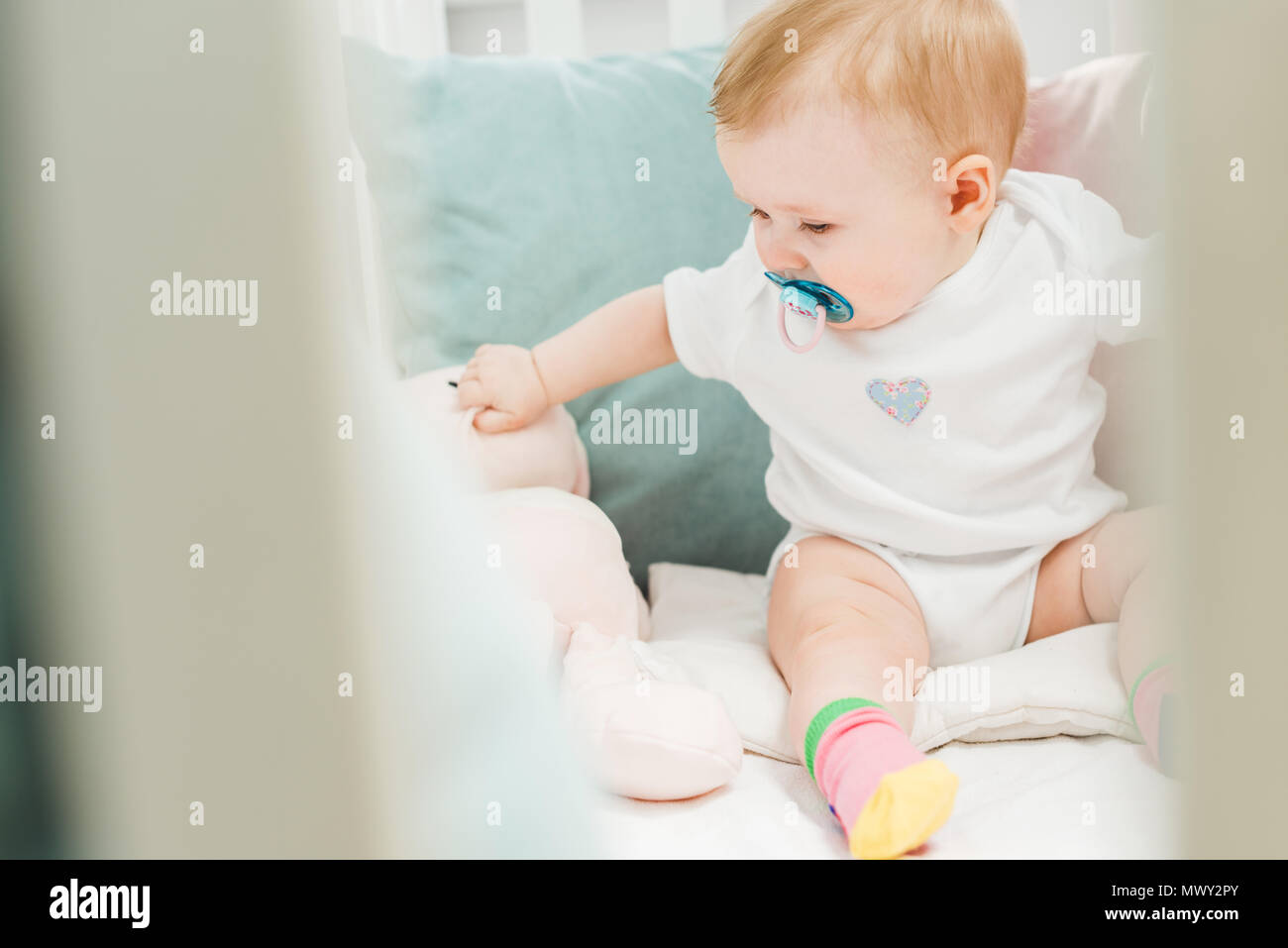 Infant with baby dummy and teddy hare sitting in crib Stock Photo