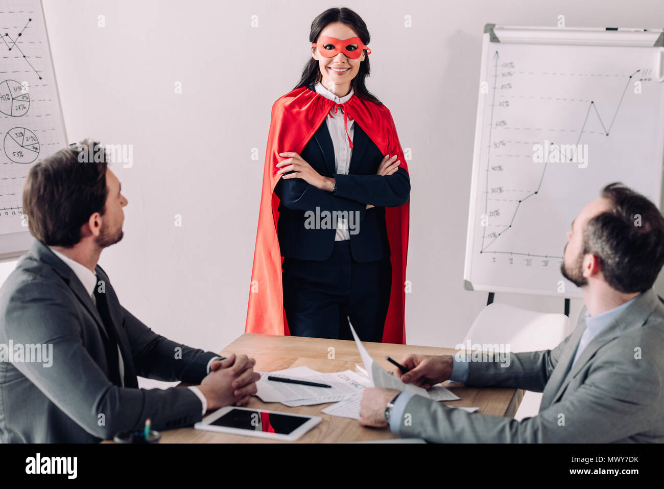smiling super businesswoman in mask and cape with businessmen in office Stock Photo