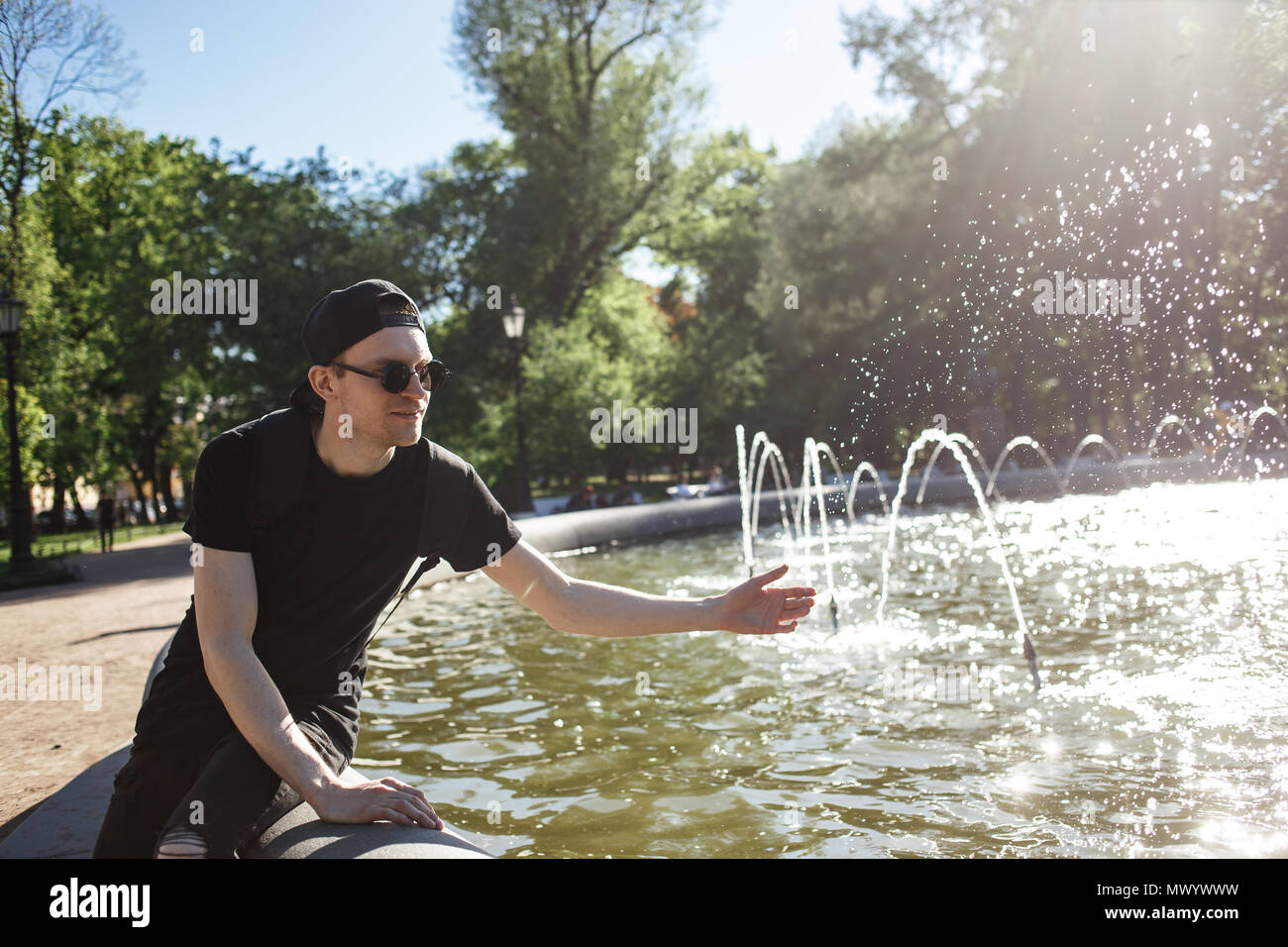 Stylish man posing at the fountain in hot summer day. He wearing in black jeans, black T-shirt, black cap with riund sunglasses Stock Photo