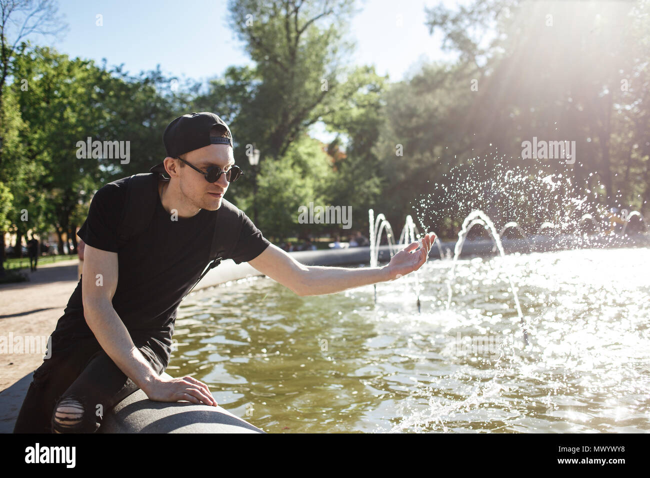 Stylish man posing at the fountain in hot summer day. He wearing in black jeans, black T-shirt, black cap with riund sunglasses Stock Photo